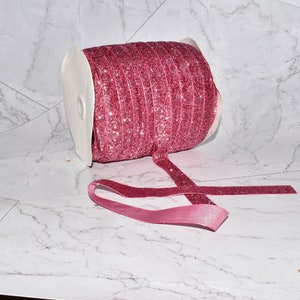 K&ouml;nnte beinhalten: Eine Rolle pinkfarbenes Glitzerband mit einer wei&szlig;en Spule. Das Band ist abgewickelt und liegt auf einer wei&szlig;en Marmorfl&auml;che.