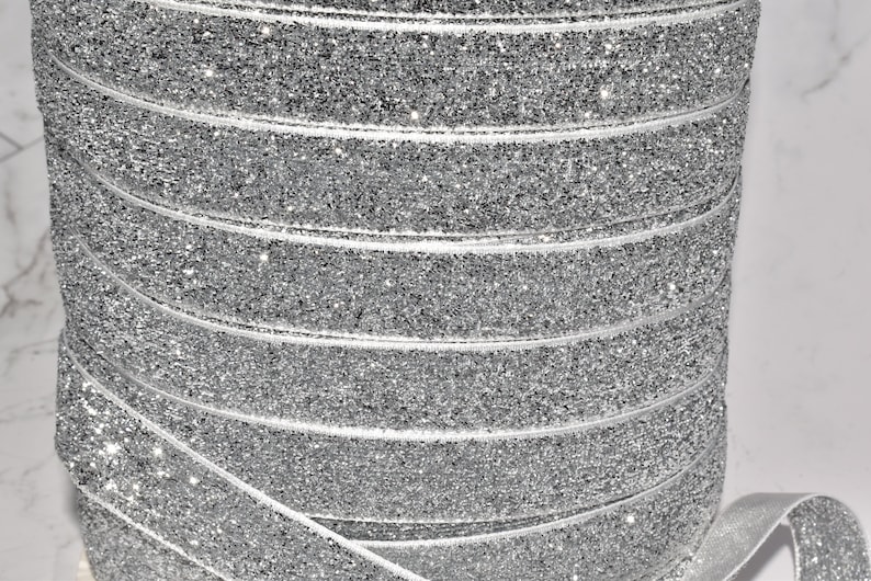 K&ouml;nnte beinhalten: Eine Rolle silbernes Glitzerband. Das Band ist aus einem weichen, dehnbaren Material gefertigt und perfekt, um jedem Projekt einen Hauch von Glanz zu verleihen.