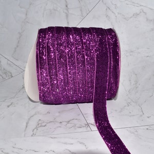 K&ouml;nnte beinhalten: Eine Rolle violettes Glitzerband mit glitzerndem Finish. Das Band ist aufgerollt und ein St&uuml;ck Band h&auml;ngt herunter.