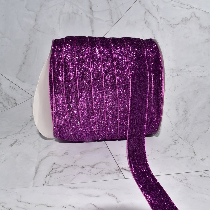 K&ouml;nnte beinhalten: Eine Rolle violettes Glitzerband mit glitzerndem Finish. Das Band ist aufgerollt und ein St&uuml;ck Band h&auml;ngt herunter.