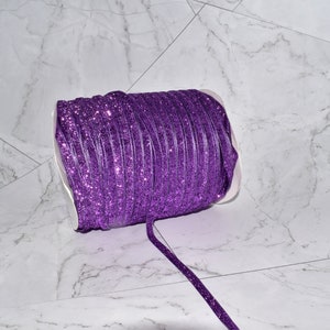 K&ouml;nnte beinhalten: Eine Rolle violettes Glitzerband. Das Band ist um einen wei&szlig;en Pappkern gewickelt und liegt auf einer wei&szlig;en Marmorfl&auml;che.