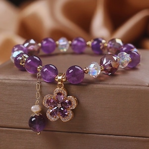May include: A purple amethyst gemstone bracelet with a gold flower charm. The bracelet is made with round amethyst beads and clear crystal beads. The flower charm has a pearl accent.
