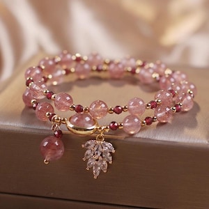 Könnte beinhalten: Ein doppelreihiges Armband mit rosa Edelsteinperlen und kleinen roten Perlen. Das Armband hat goldfarbene Akzente und zwei Anhänger: ein runder rosa Stein und ein blattförmiger Anhänger mit klaren Kristallen. Das Armband wird auf einer goldfarbenen Oberfläche präsentiert.