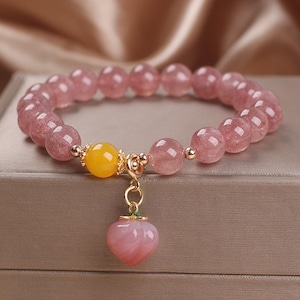 May include: A pink quartz bracelet with a yellow and pink charm in the shape of a peach. The bracelet is made of small, round beads.