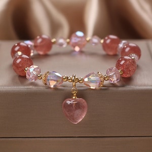 May include: A pink crystal bracelet with a heart-shaped charm. The bracelet is made with round and faceted beads, and the charm is made of a clear crystal with a pink hue.