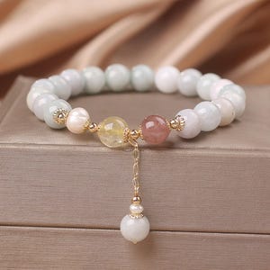 以下が含まれることがあります： 薄い青色の宝石のブレスレットで、金色のチェーンとチャームが付いています。ブレスレットには、真珠、黄色の宝石、ピンク色の宝石が付いています。