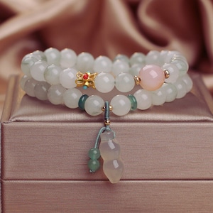 Op de afbeelding: Twee jade armbanden met een roze jade kraal en een gouden bedel. De armbanden liggen op een lichtbruine doos met een groene jade hanger.
