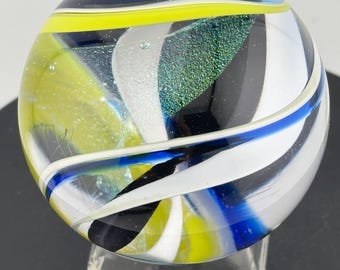 Pode incluir: Uma bola de gude de vidro transparente com um design colorido. A bola de gude apresenta redemoinhos brancos, pretos, azuis e amarelos. Uma seção da bola de gude tem uma aparência verde iridescente texturizada. Ela repousa em um suporte hexagonal transparente.