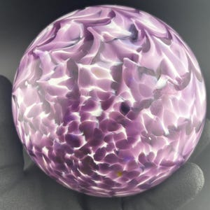 May include: A spherical glass art piece with a swirling pattern of lavender and white. The orb is held in a gloved hand, showcasing the intricate design and color variations. The glass appears to have a speckled texture.