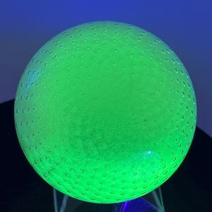 May include: A glowing green glass sphere with a textured surface, resting on a clear, geometric stand. The sphere is illuminated, creating a vibrant, luminous effect. The sphere has a series of small, evenly spaced dimples across its surface.