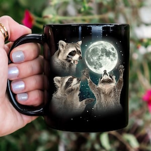 May include: Black coffee mug featuring a white design of three raccoons looking up at a full moon. The raccoons are in a playful pose, with one reaching up to touch the moon.