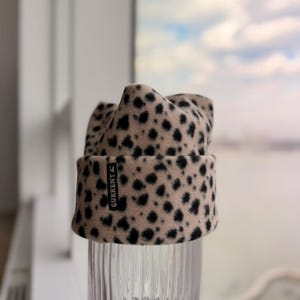 May include: A beige beanie with black spots, resembling a leopard print. The hat has a folded brim and a small black tag that reads "CURRENT". The hat is displayed on a clear, ribbed glass.