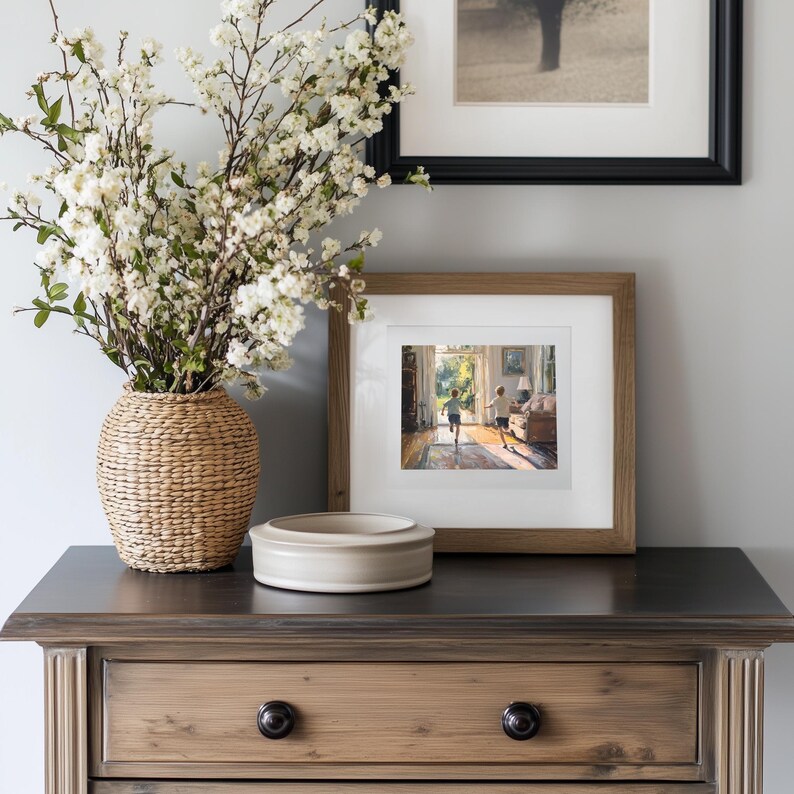 May include: A wooden chest of drawers with a drawer and two knobs. A white ceramic bowl and a woven basket with white flowers are on top of the dresser. A framed picture of two children running towards a doorway is on the dresser.