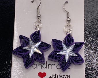 Può includere: Un paio di orecchini a forma di fiore viola fatti a mano con centri a stella argentati. Gli orecchini sono su una carta bianca con le parole "handmade with love" e un cuore rosso.