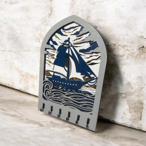 May include: A grey wooden key holder featuring a navy blue and white sailboat design. The key holder has six black hooks and a decorative cutout of waves and clouds. The key holder is approximately 30 cm in height.