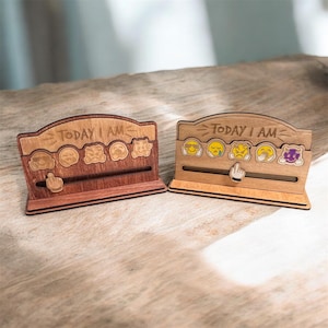 Puede incluir: Dos organizadores de escritorio de madera con el texto "TODAY I AM" encima de una fila de caras emoji. Cada organizador tiene un puntero deslizante con un icono de mano. Uno está teñido de un marrón rojizo oscuro, el otro es de color claro.