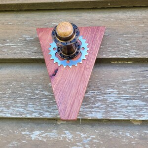 May include: A triangular wooden wall art piece with a bicycle gear and hub assembly. The wood is reddish-brown, and the gear is silver. A cork-like material tops the hub. The piece is mounted on a weathered, light-colored wooden surface.