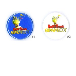 May include: Two circular buttons featuring the text "Monty Python's SPAMALOT". One button has a blue background, the other is white. Both buttons have a gold chalice design with flags.