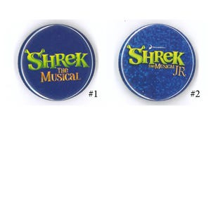May include: Two round, blue buttons featuring the text "Shrek The Musical" and "Shrek The Musical JR" in green and yellow. The buttons have a white border and are set against a white background.