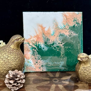 May include: Abstract painting on a square canvas with green, white, and orange colours. The painting has a swirling, fluid design with gold glitter accents.