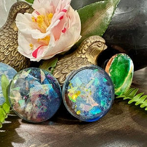 May include: Four round, iridescent resin bottle stoppers with a glittery, confetti-like design. The stoppers are displayed on a wooden surface with a green fern and gold bird figurines.