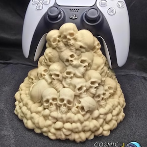 May include: A white and black PlayStation 5 controller rests atop a beige sculpture of numerous skulls. The sculpture has a textured, organic appearance. The image includes the text "COSMIC MANATEE PRINTING" and a cartoon astronaut.