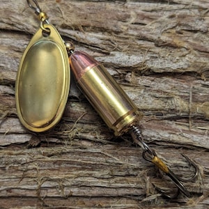 May include: A fishing lure with a gold-colored, teardrop-shaped spinner blade and a brass bullet casing. The lure has a silver hook and is set against a textured, brown wooden background.