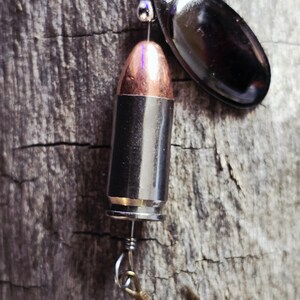 May include: A fishing lure featuring a bullet casing with a copper tip, a black metal blade, and a gold-colored hook. The lure is suspended by a silver wire and is set against a weathered wood background.