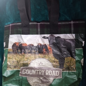 Könnte beinhalten: Eine wiederverwendbare Tragetasche mit schwarzen Griffen und einem grün-schwarz karierten Oberteil. Der Hauptteil der Tasche zeigt eine Grafik einer Rinderherde auf einem Feld mit den Worten "COUNTRY ROAD" und "CALE".