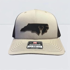 May include: A tan and black trucker hat with a black leather patch featuring the outline of North Carolina and a deer head. The hat has a curved brim and an adjustable strap. The Richardson 112 Trucker hat is adjustable.