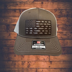 May include: A gray and white trucker hat with a black and white American flag design featuring a silhouette of fish. The hat has a white mesh back and a black label with the text "EST. 1970 RICHARDSON. ORIGINAL 112 TRUCKER ADJUSTABLE".