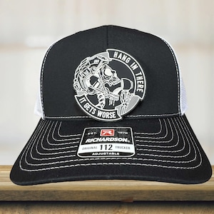 May include: Black and white trucker hat with a patch featuring a skull wearing a football helmet and the text "Hang in There, It Gets Worse." The hat has a white mesh back and a curved brim. The Richardson brand label is visible.