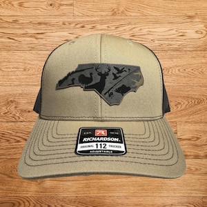 May include: Tan and black trucker hat with a patch featuring a silhouette of North Carolina with hunting and wildlife imagery. The hat has a curved brim and a mesh back. The patch includes a deer, ducks, and a fish. The label reads "RICHARDSON ORIGINAL 112 TRUCKER ADJUSTABLE".
