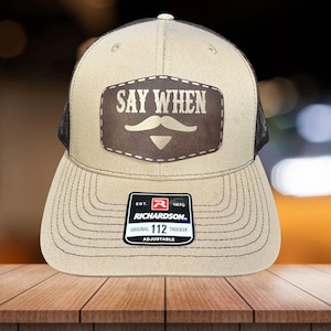 May include: Tan and black trucker hat with the text "SAY WHEN" above a mustache graphic. The hat has a curved brim and an adjustable strap. The Richardson brand label is visible.