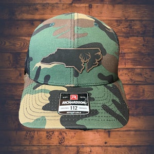 May include: Camouflage trucker hat with a black mesh back and a curved brim. The hat features a patch with the outline of North Carolina and a deer head. The Richardson brand label is visible.
