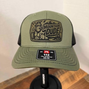 May include: An olive green and black trucker hat featuring a rectangular patch with a graphic of a Bigfoot character wearing sunglasses and a hat, holding a beverage, with the text MOUNTAIN DUDE. A black sticker on the brim reads R 112 The Original MD-LG.