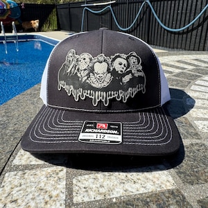Peut inclure: Casquette de camionneur noire et blanche avec un motif de personnages de films d'horreur. La casquette a une visière et une couronne noires avec un dos en filet blanc. La casquette Richardson 112 est réglable.