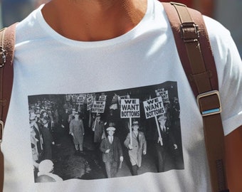 May include: A white t-shirt with a black and white photo of a group of people holding signs that say "We Want Bottoms". The photo is from a protest march. The person wearing the shirt is standing in a crowd of people.