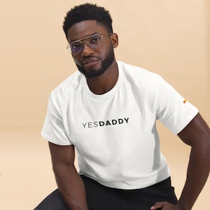 May include: A white t-shirt with the text "YESDADDY" printed in black on the front. The shirt has a rainbow flag sewn on the right sleeve.