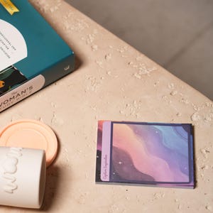 Puede incluir: Un libro verde azulado y dorado titulado "The Woman's Study Bible" es parcialmente visible. También hay un pequeño bloc de notas adhesivas coloridas con un diseño ondulado y el texto "Capture Inspiration", junto con una taza blanca y un posavasos color melocotón.