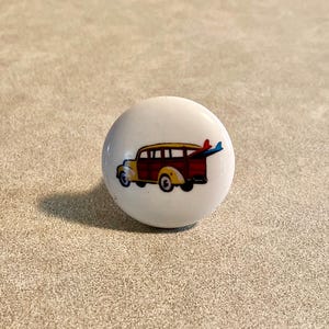 May include: A white ceramic cabinet knob featuring a colorful illustration of a vintage woodie station wagon with surfboards. The car is red, yellow, and brown, with blue surfboards attached to the roof. The knob is round and has a glossy finish.