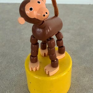 May include: A wooden monkey toy with a brown body, head, and limbs. The monkey has a tan face and feet, and a brown tail. It stands on a yellow cylinder base. The toy is likely a vintage or retro item.