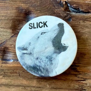 May include: Round button with a howling wolf illustration in black and white. The word "SLICK" is printed in black above the wolf. The button is set against a wooden background.