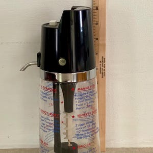 May include: Vintage cocktail shaker with a black top and a clear glass base. The glass is printed with cocktail recipes. A wooden ruler is placed next to the shaker, measuring up to 12 inches.