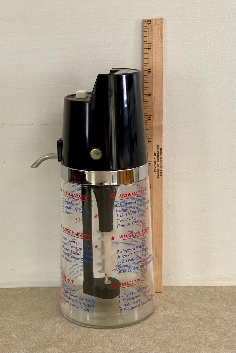 May include: Vintage cocktail shaker with a black top and a clear glass base. The glass is printed with cocktail recipes. A wooden ruler is placed next to the shaker, measuring up to 12 inches.