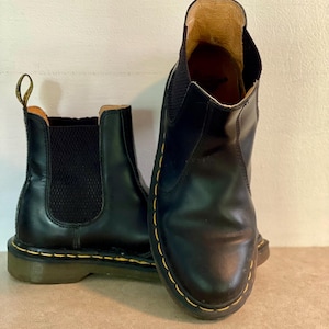 May include: A pair of black leather Chelsea boots with elastic side panels. The boots feature yellow stitching around the soles and a pull tab at the back. The boots are a classic style.