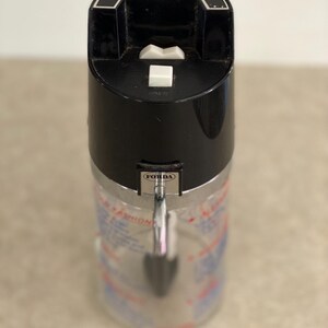May include: A vintage Drinko-Matic measure-pourer with a black top and a silver-colored body. The top features the text "DRINKO-MATIC MEASURE-POURER" and a white button. The body has measurement markings.