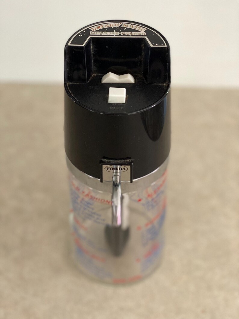 May include: A vintage Drinko-Matic measure-pourer with a black top and a silver-colored body. The top features the text "DRINKO-MATIC MEASURE-POURER" and a white button. The body has measurement markings.