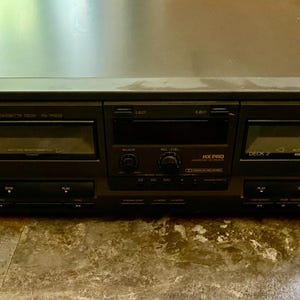May include: A black Technics stereo cassette deck. The deck has two cassette slots, a display screen, and various control buttons. The Technics logo is visible on the top left. The unit is sitting on a patterned surface.