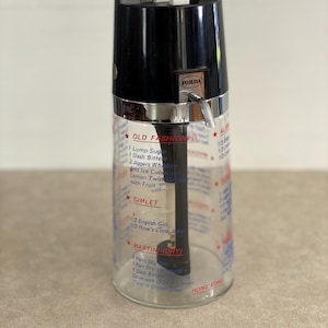 May include: Vintage cocktail shaker with a black top and a clear glass body. The shaker has a silver band and a spout. The glass is printed with cocktail recipes, including "Old Fashioned" and "Gimlet". The brand name "FORDA" is visible.